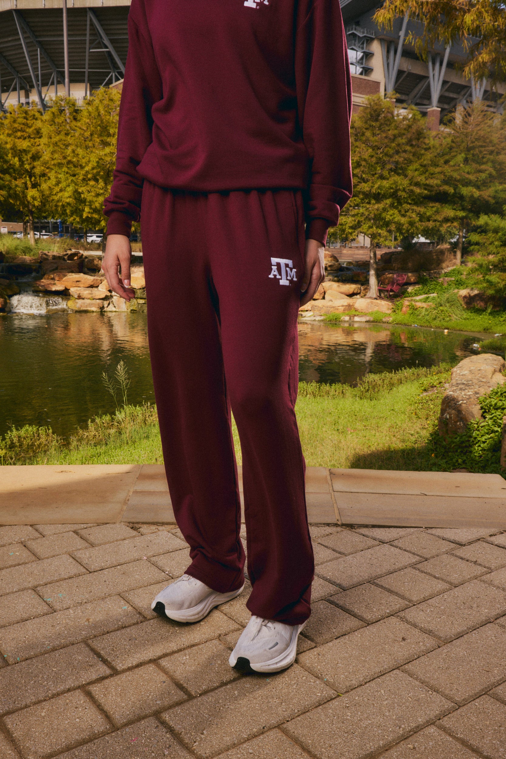 A person wearing a matching maroon sweatshirt and sweatpants set with white sneakers, standing outdoors on a paved walkway with a pond, trees, and stadium seating in the background. The sweatpants have a white 'ATK' logo on the left thigh.