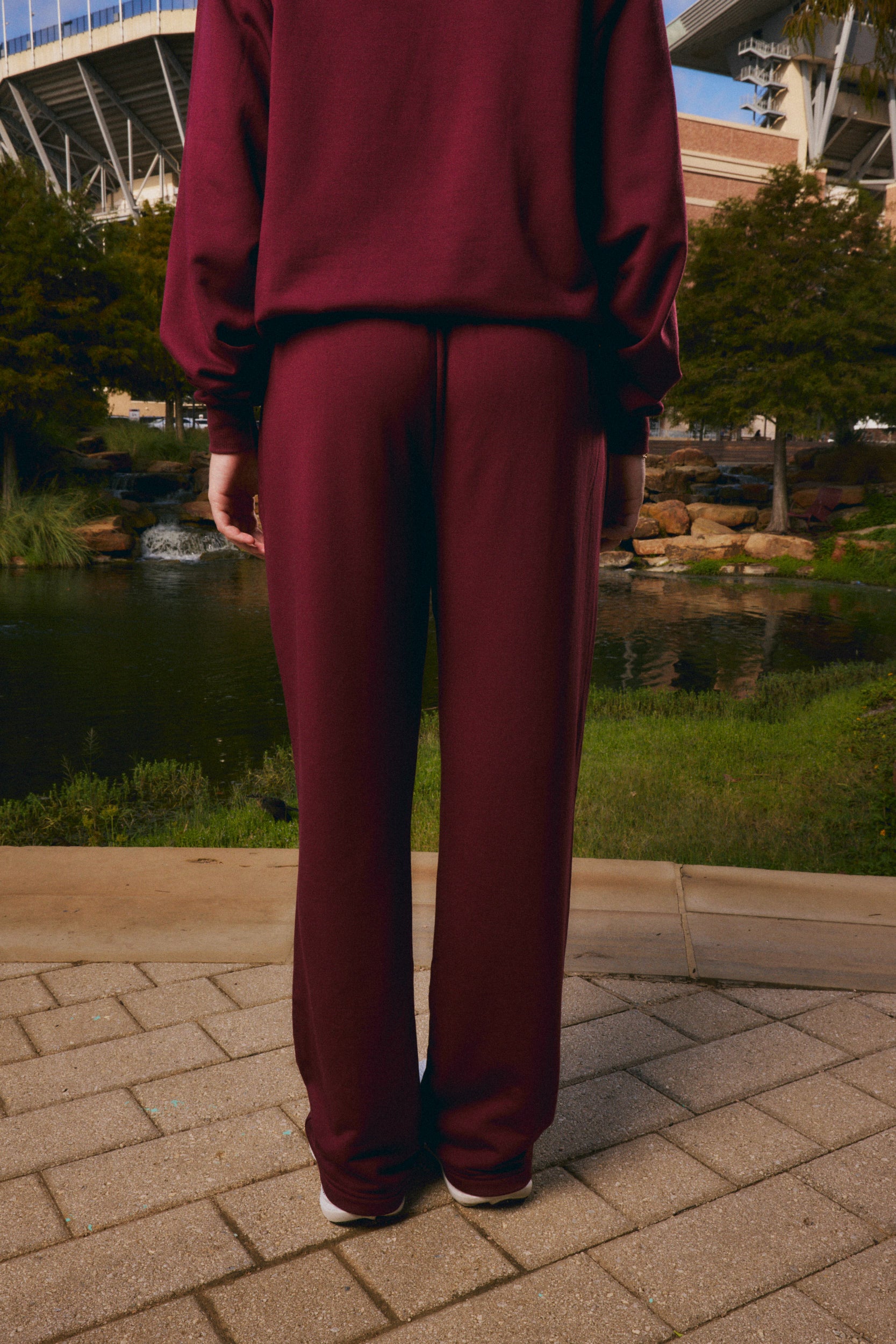 A person viewed from behind wearing a matching burgundy sweatshirt and wide-leg sweatpants, standing on a paved walkway outdoors near a pond and trees.