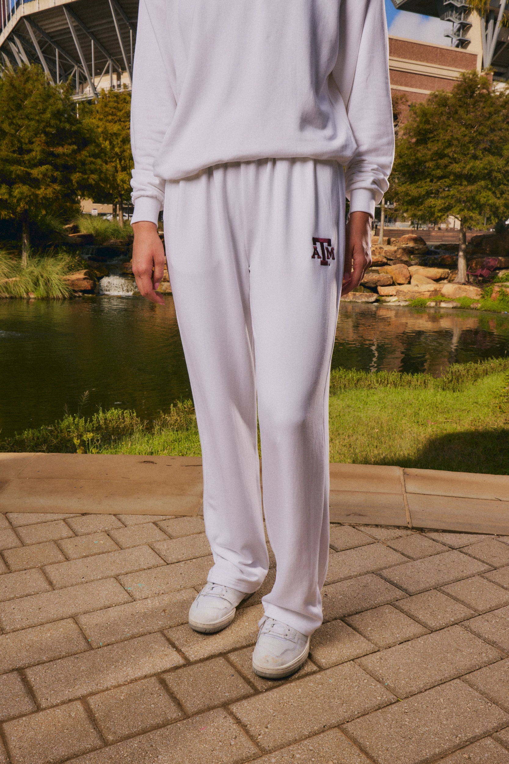 A person wearing a white sweatshirt and white sweatpants with a maroon 'AF' logo on the left thigh, standing outdoors on a paved walkway near a pond and trees.