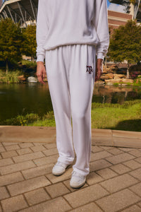 A person wearing a white sweatshirt and white sweatpants with a maroon 'AF' logo on the left thigh, standing outdoors on a paved walkway near a pond and trees.