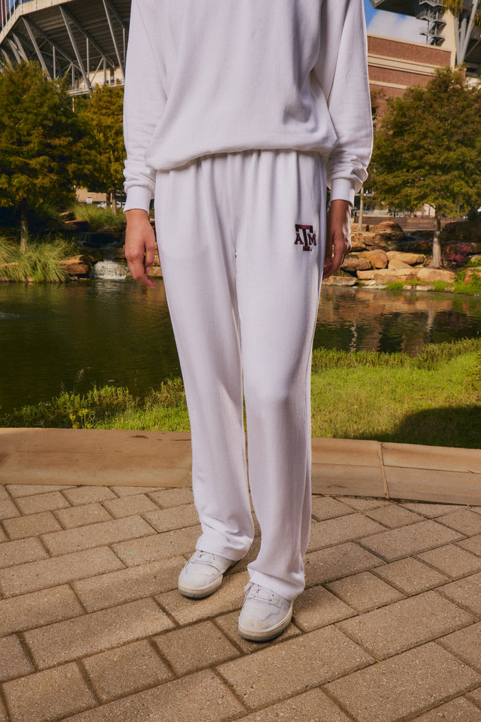 A person wearing a white sweatshirt and white sweatpants with a maroon 'AF' logo on the left thigh, standing outdoors on a paved walkway near a pond and trees.