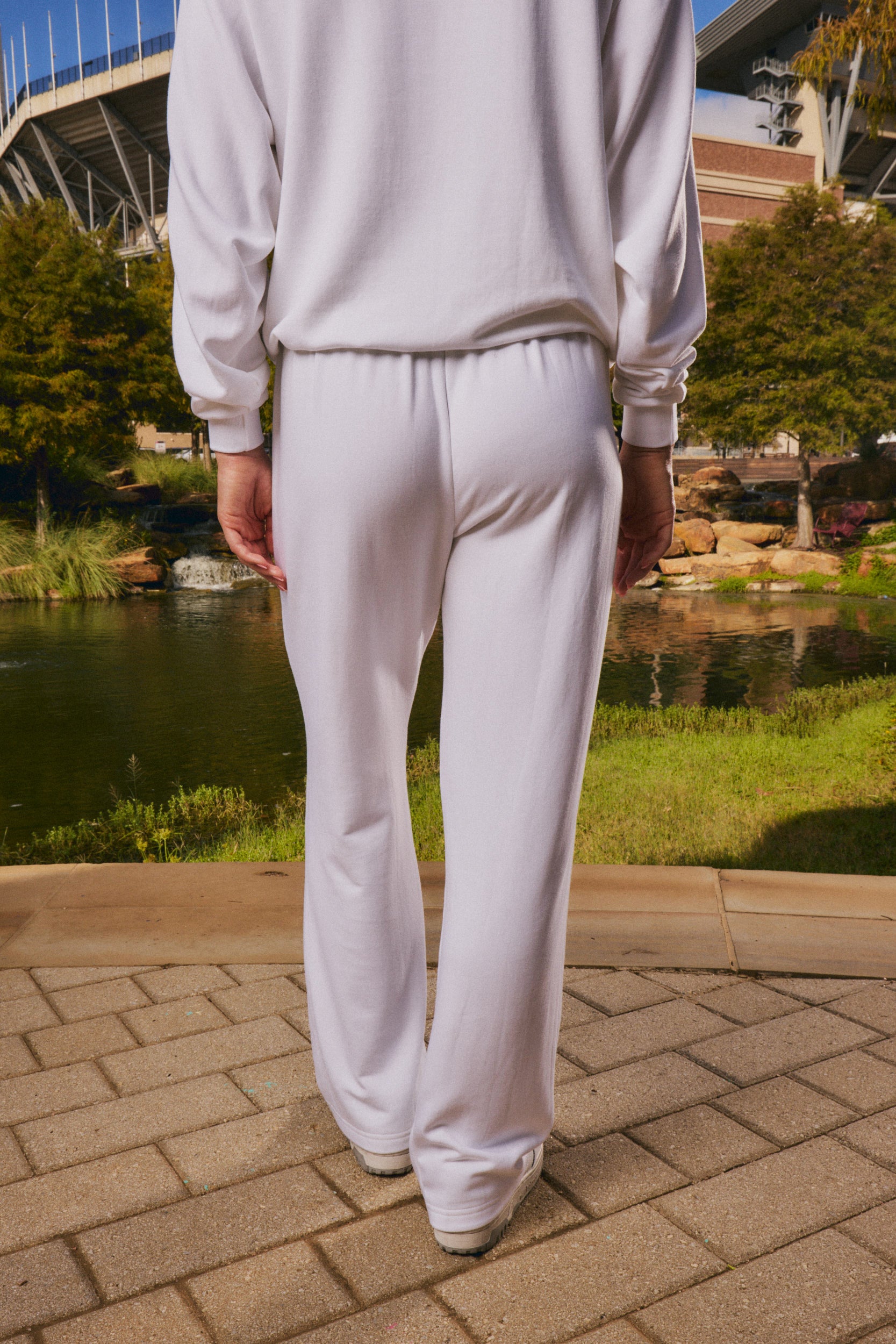 A person viewed from behind wearing a matching white sweatshirt and wide-leg sweatpants, standing on a paved walkway outdoors near a pond and greenery.