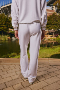 A person viewed from behind wearing a matching white sweatshirt and wide-leg sweatpants, standing on a paved walkway outdoors near a pond and greenery.