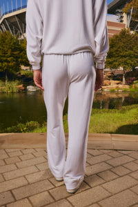 A person viewed from behind wearing a matching white sweatshirt and wide-leg sweatpants, standing on a paved walkway outdoors near a pond and greenery.