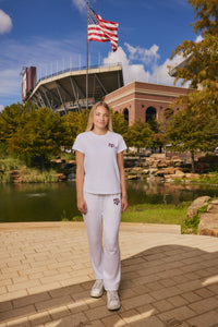 A woman stands outdoors in front of a stadium and pond, wearing a matching white athletic set with a small red Texas Tech logo on the chest and left thigh.