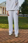 A person wearing a white sweatshirt and matching sweatpants stands outdoors on a brick path with grass and a classical building in the background. The sweatpants have a small navy graphic on the left thigh.