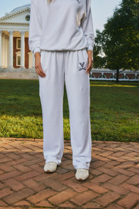 A person wearing a white sweatshirt and matching sweatpants stands outdoors on a brick path with grass and a classical building in the background. The sweatpants have a small navy graphic on the left thigh.