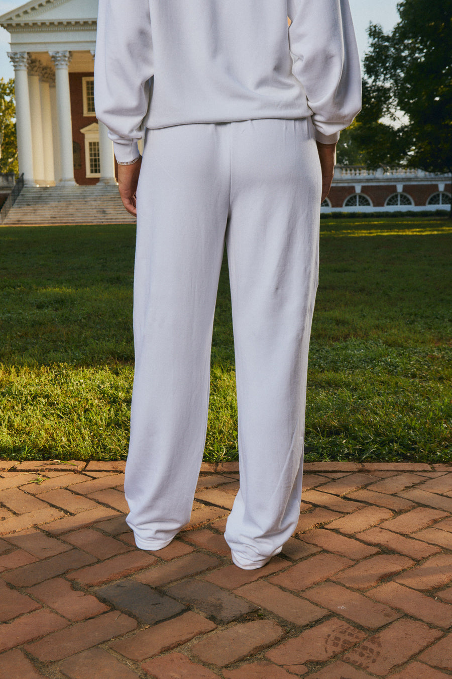 A person wearing a matching white sweatshirt and wide-leg sweatpants, shown from the back in an outdoor setting with a brick path, green lawn, and a classical building in the background.