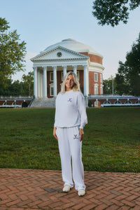 A woman stands outdoors in front of a large classical building on a grassy lawn, wearing a white long-sleeve top and matching white pants with small dark embroidered logos.