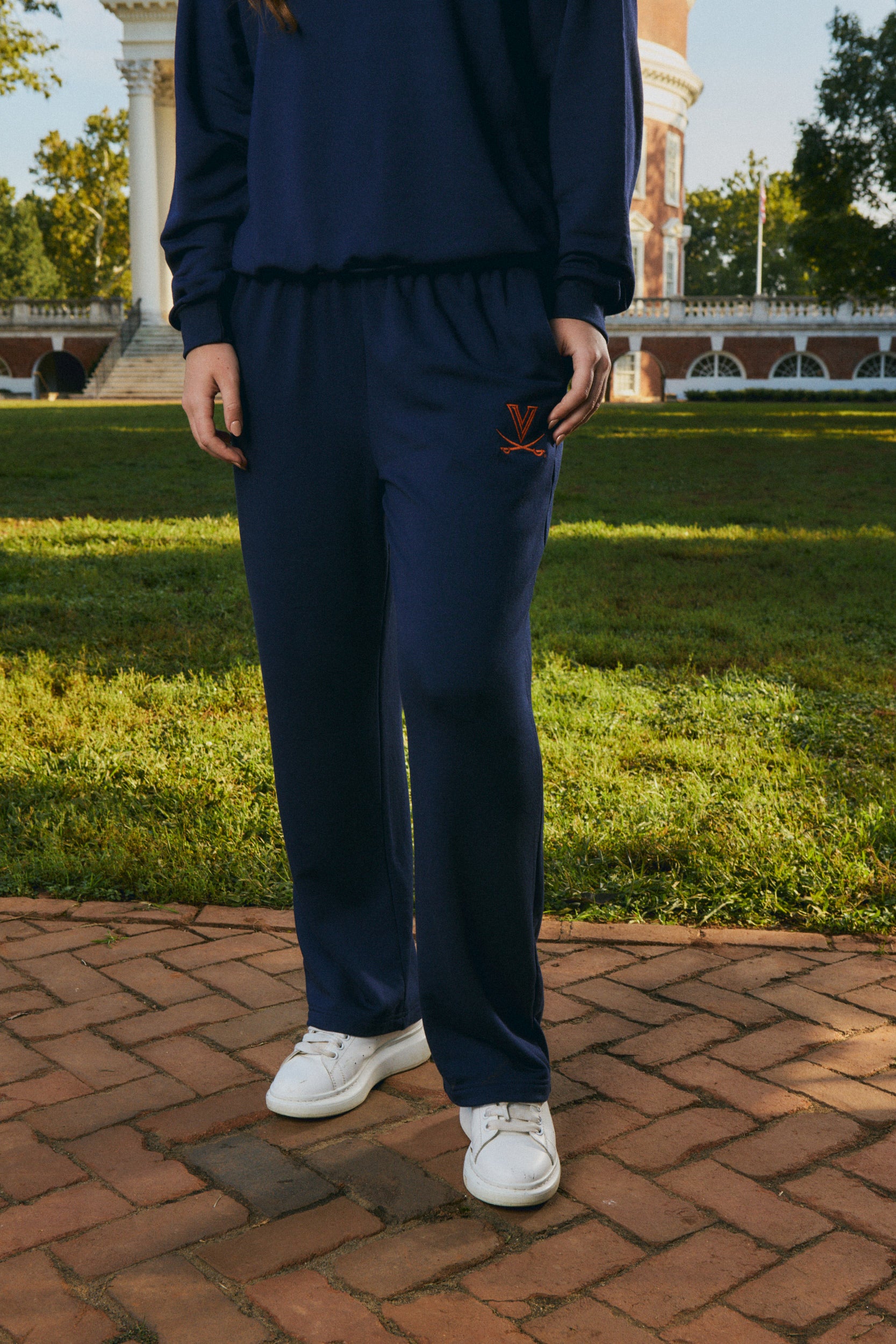 A person wearing a navy blue sweatshirt and matching sweatpants with a small orange embroidered logo on the left thigh, standing outdoors on a brick path with grass and a building in the background.