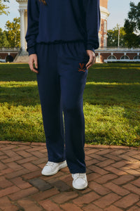 A person wearing a navy blue sweatshirt and matching sweatpants with a small orange embroidered logo on the left thigh, standing outdoors on a brick path with grass and a building in the background.