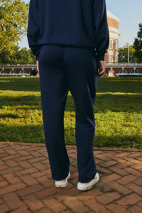 A person viewed from behind wearing a navy blue sweatshirt and matching sweatpants, standing on a brick path in a park with a large brick building and green lawn in the background.