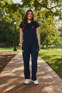 A woman stands on a brick pathway in a park wearing a navy blue short-sleeve top and matching sweatpants with a small red logo on the chest and thigh, paired with white sneakers.