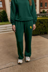 A person wearing a dark green sweatshirt and matching sweatpants with a yellow 'BU' logo on the left thigh, standing outdoors on a concrete walkway in front of a brick building.