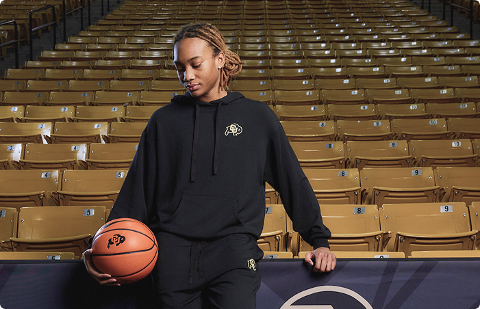 A woman stands in an indoor arena holding a basketball, wearing a black hoodie and matching sweatpants with a small gold and white logo on the chest and thigh. The background shows rows of empty tan stadium seats.
