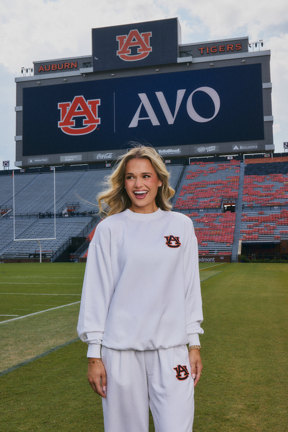 A woman stands on a football field in a stadium wearing a white Auburn Tigers matching sweatshirt and sweatpants set with a brown and orange logo. Behind her is a large scoreboard with Auburn branding.
