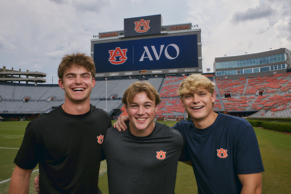 Three young men smiling and posing together on a football field in front of a large stadium scoreboard with Auburn University branding.