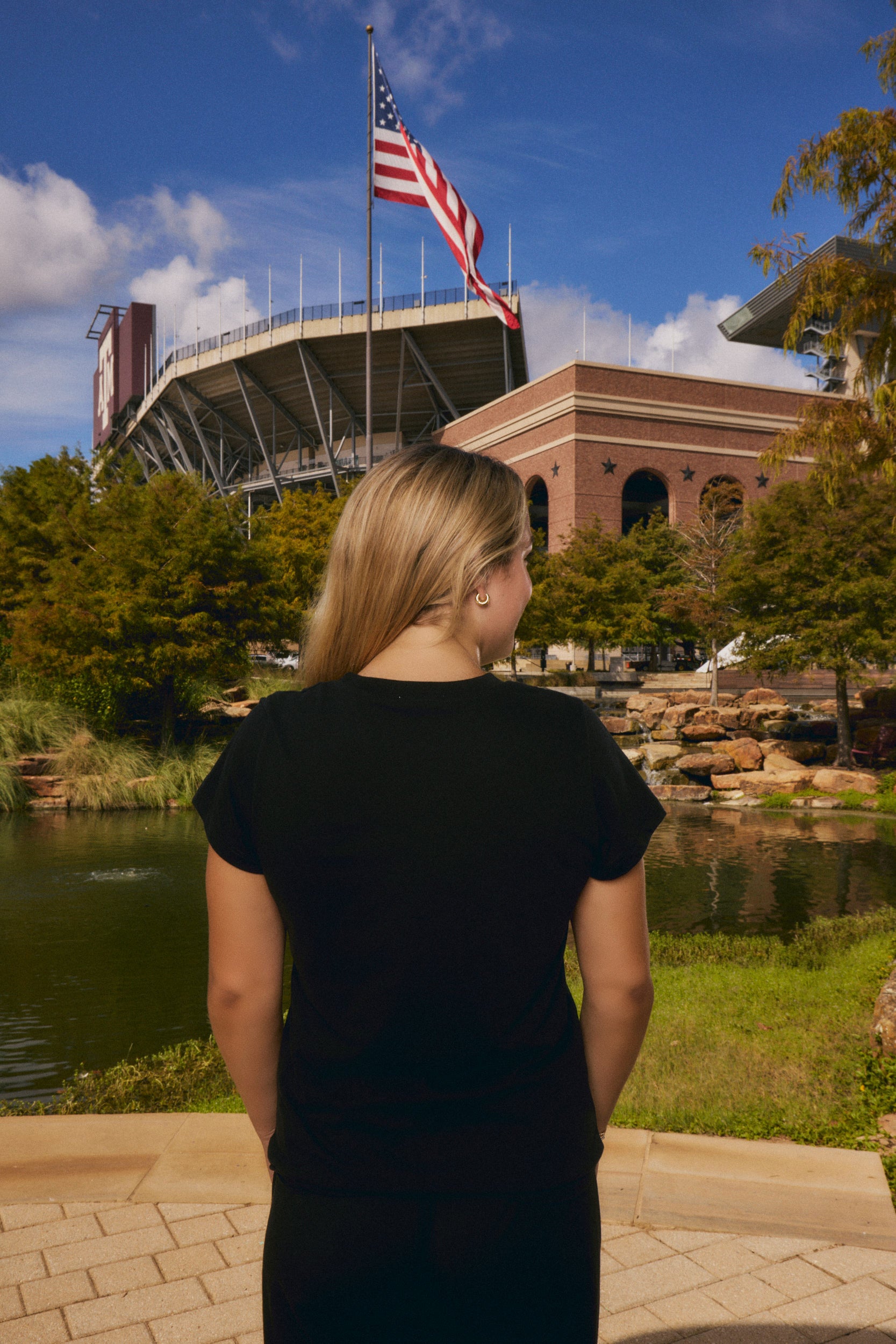 A woman with blonde hair, seen from behind, wearing a black t-shirt and black pants, standing outdoors in front of a stadium and lake with an American flag flying high under a blue sky.