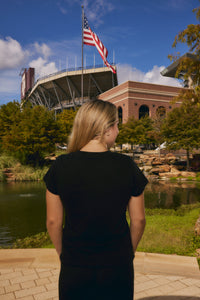 A woman with blonde hair, seen from behind, wearing a black t-shirt and black pants, standing outdoors in front of a stadium and lake with an American flag flying high under a blue sky.