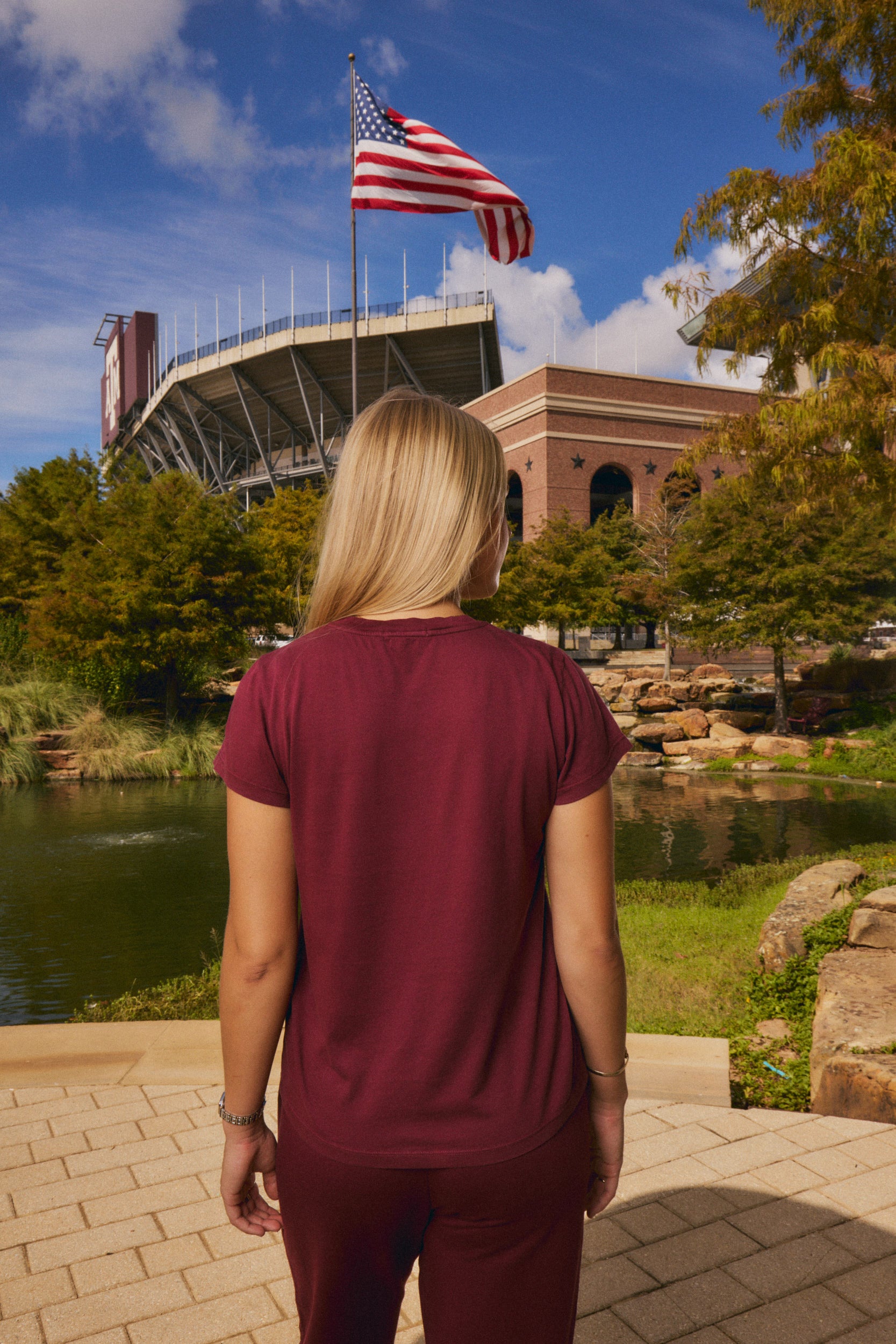 A woman seen from behind wearing a maroon short-sleeve athletic t-shirt and matching maroon pants, standing outdoors in front of a stadium with an American flag, trees, a pond, and a brick building under a blue sky.
