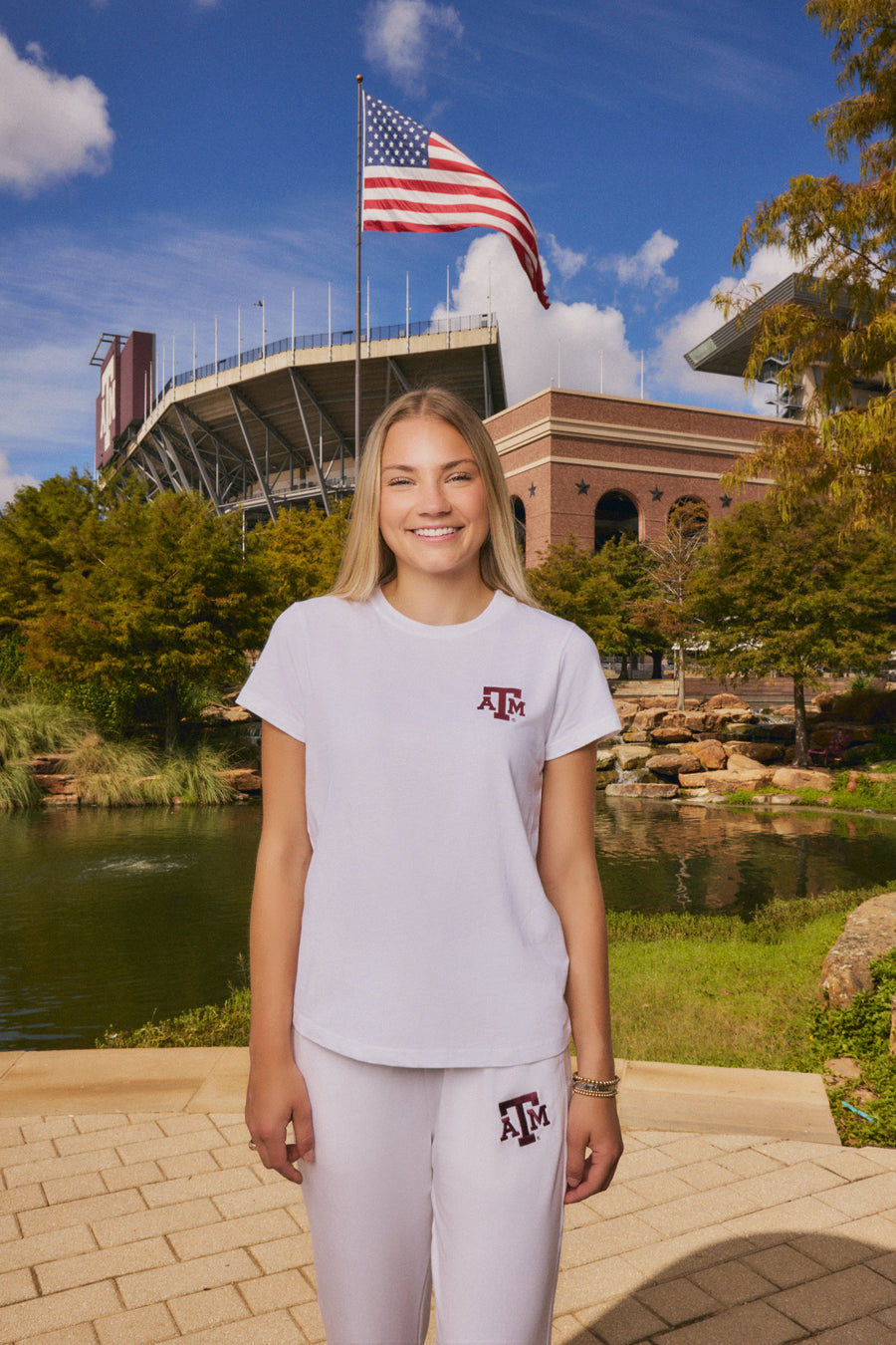 A smiling young woman stands outdoors in front of a stadium and pond, wearing a white short-sleeve T-shirt and matching sweatpants with a maroon Texas A&M logo on the chest and thigh.