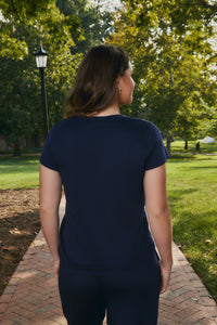 A woman viewed from behind wearing a dark navy blue short-sleeve t-shirt and matching navy pants, standing on a brick pathway in a sunny park with green trees in the background.