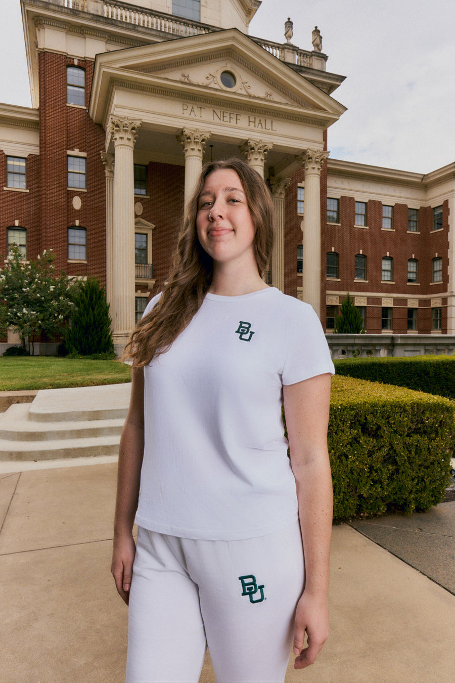 A woman stands outdoors in front of a large classical brick building, wearing a white short-sleeve t-shirt and matching white sweatpants with a green 'BU' logo on the chest and thigh.