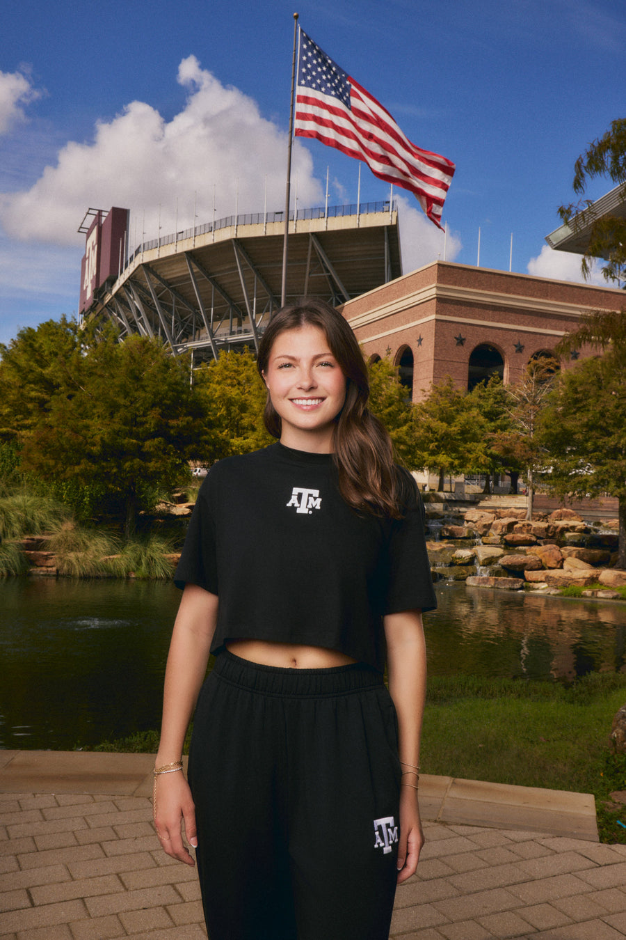 A smiling young woman stands outdoors in front of a stadium and pond, wearing a black cropped T-shirt and black sweatpants with white Texas A&M logo graphics. An American flag flies above, with trees, water, and a brick building in the background.