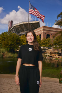 A smiling young woman stands outdoors in front of a stadium and pond, wearing a black cropped T-shirt and black sweatpants with white Texas A&M logo graphics. An American flag flies above, with trees, water, and a brick building in the background.