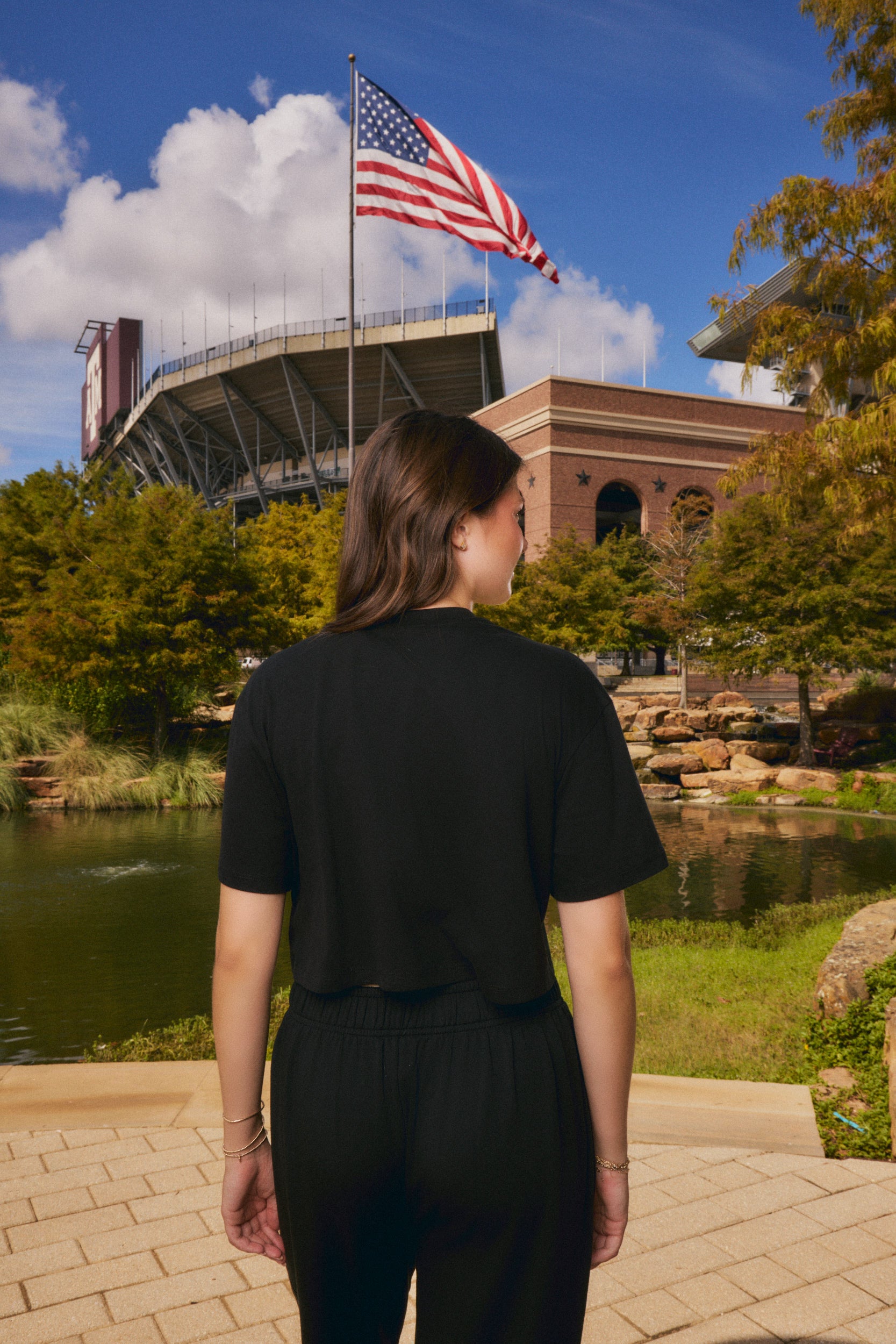 A woman viewed from behind wearing a black short-sleeve crop top and black pants, standing outdoors in front of a stadium and an American flag, with trees, a pond, and a brick building in the background.