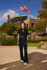 A woman stands outdoors in front of a stadium and lake, wearing a matching black cropped T-shirt and sweatpants set with white logos. An American flag is visible in the background, along with trees, rocks, and a brick building.