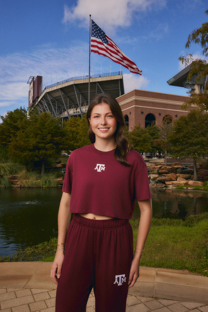 A woman stands outdoors in front of a stadium and pond, wearing a matching maroon cropped T-shirt and sweatpants set with a white Texas A&M logo. An American flag is visible in the background.