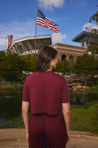 A woman viewed from behind wearing a matching burgundy short-sleeve crop top and pants set, standing outdoors in front of a stadium, an American flag, trees, and a pond.