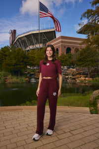 A woman stands outdoors in front of a stadium and pond wearing a matching maroon athletic crop top and sweatpants set with white sneakers. The apparel features a white Texas A&M logo.