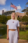 A smiling blonde woman stands outdoors in front of a stadium and pond, wearing a white cropped T-shirt and matching sweatpants with a maroon Texas A&M logo on the chest and thigh.