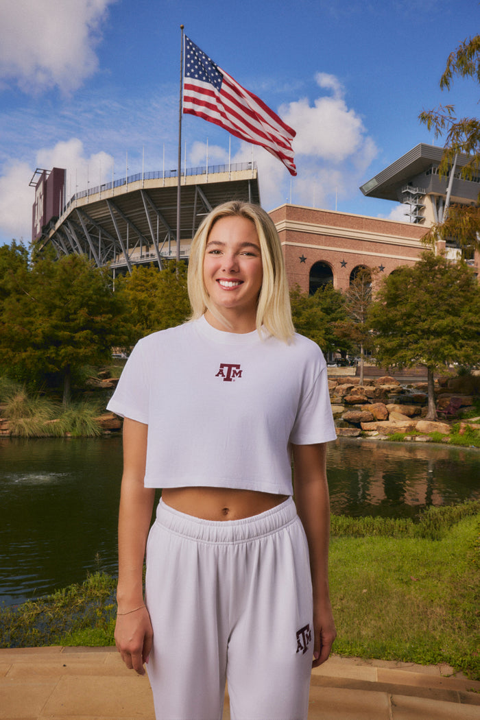 A smiling blonde woman stands outdoors in front of a stadium and pond, wearing a white cropped T-shirt and matching sweatpants with a maroon Texas A&M logo on the chest and thigh.