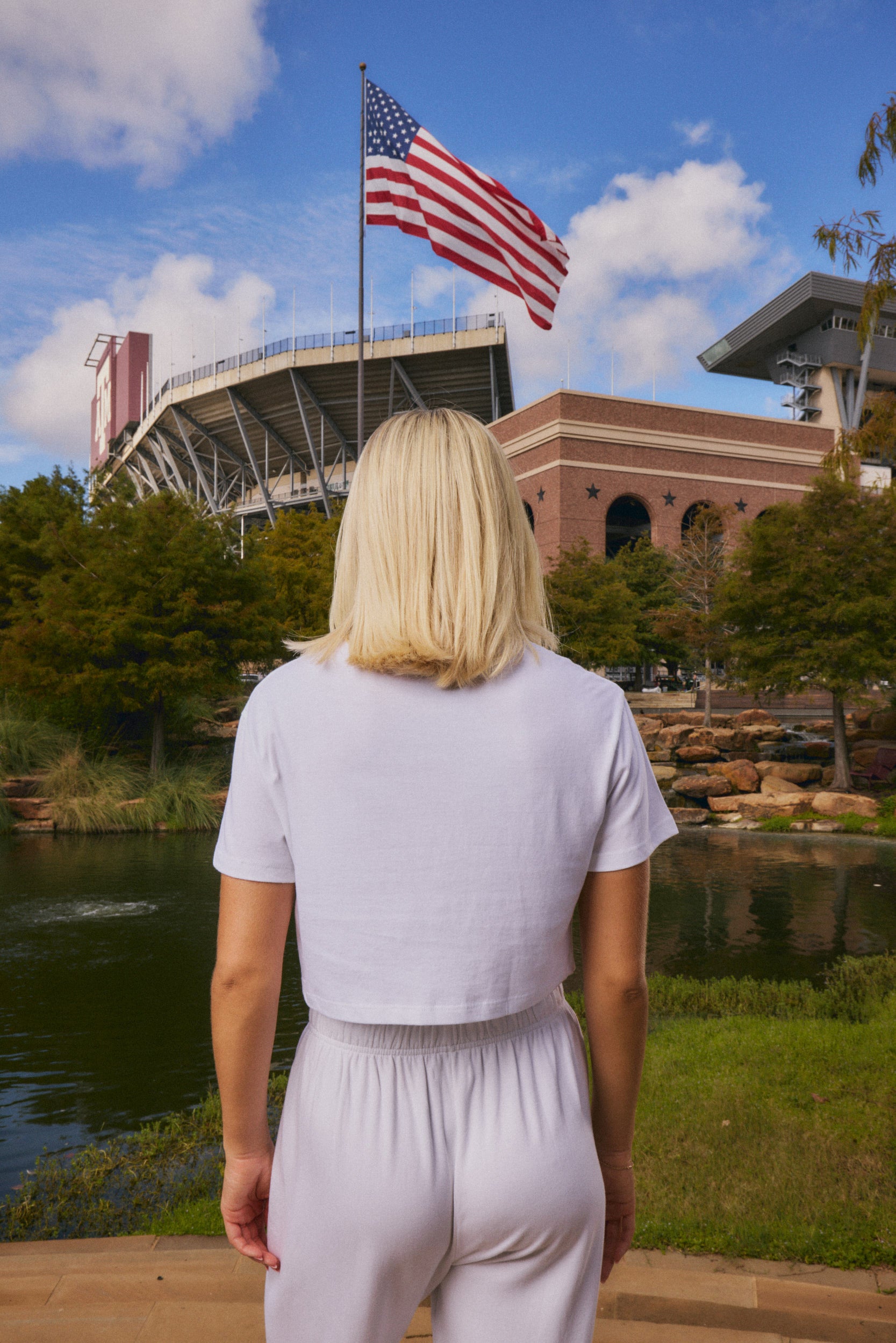 A blonde woman seen from behind wearing a white short-sleeve crop top and white sweatpants, standing outdoors in front of a stadium, an American flag, and a pond.