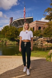 A young woman stands outdoors in front of a stadium and pond, wearing a white cropped T-shirt with a small red logo, black sweatpants, and white sneakers.