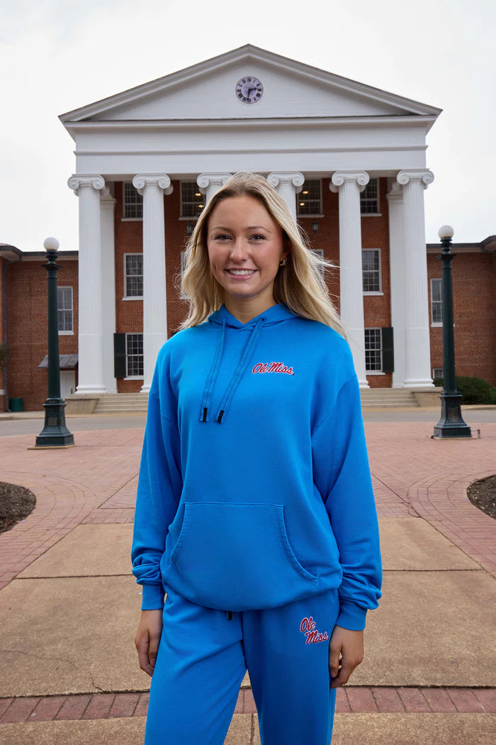 Woman wearing a bright blue Ole Miss hoodie and matching sweatpants with red embroidered logo details, posing in front of a university building with white columns. The relaxed-fit collegiate lounge set features a drawstring hood, kangaroo pocket, and coordinated campus-inspired style.