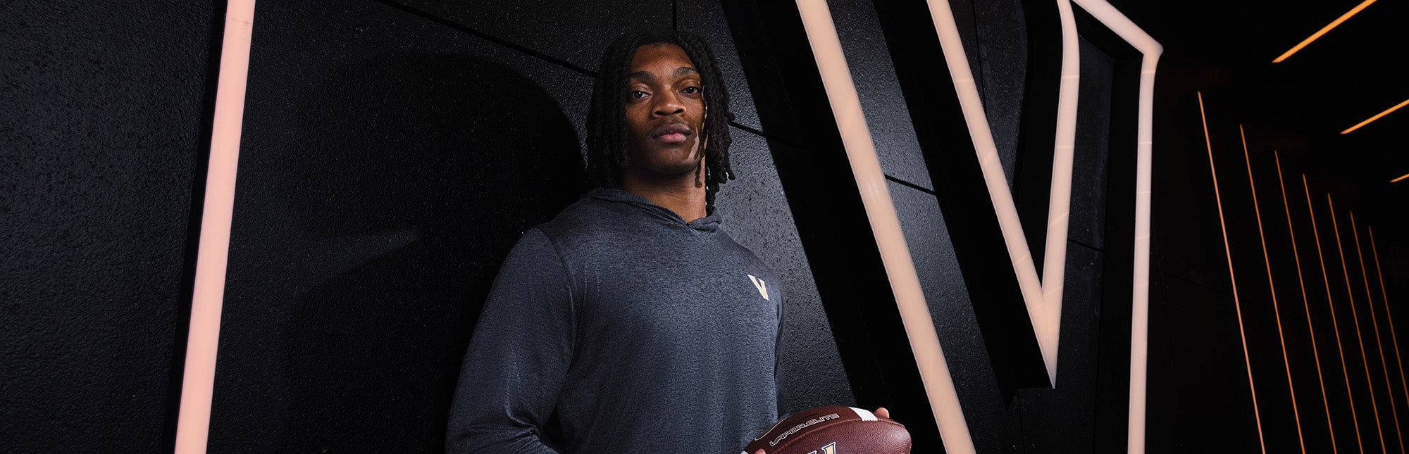 Vanderbilt football player holding a football, standing against a dark modern wall with vertical glowing light strips, wearing a gray performance hoodie with a Vanderbilt “V” logo.