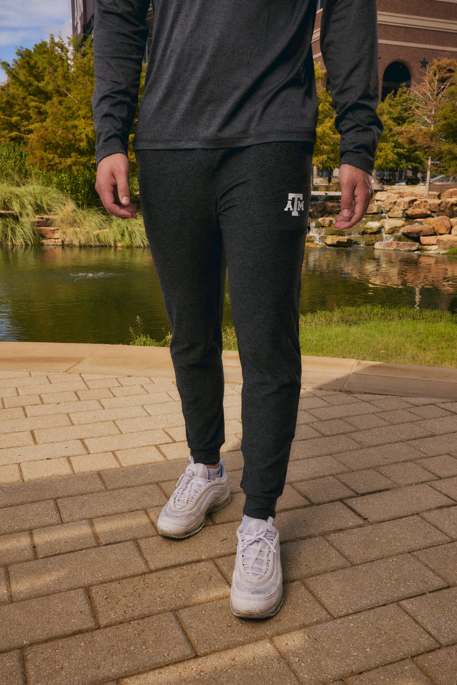 A person outdoors wearing dark charcoal heather jogger sweatpants with a white 'JFC' logo on the left thigh, paired with a matching dark long-sleeve top and white sneakers, standing on a paved walkway near a pond.