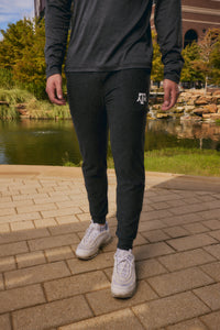 A person outdoors wearing dark charcoal heather jogger sweatpants with a white 'JFC' logo on the left thigh, paired with a matching dark long-sleeve top and white sneakers, standing on a paved walkway near a pond.