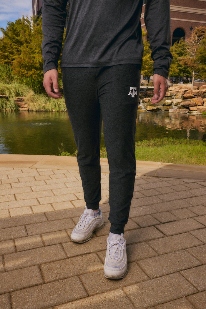 A person outdoors wearing dark charcoal heather jogger sweatpants with a white 'JFC' logo on the left thigh, paired with a matching dark long-sleeve top and white sneakers, standing on a paved walkway near a pond.