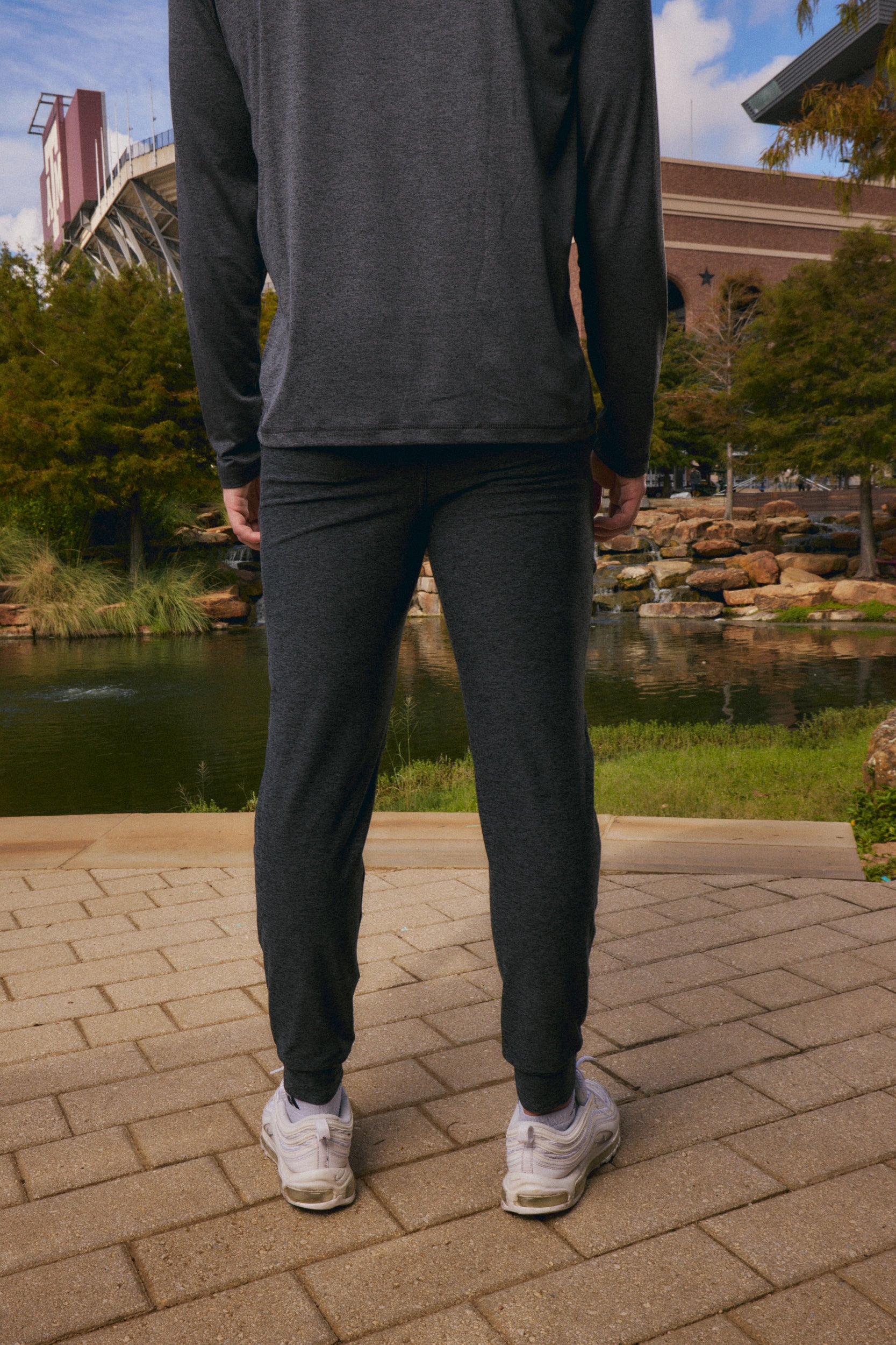 A person viewed from behind wearing dark charcoal gray athletic jogger pants and a matching long-sleeve top, standing on a paved walkway outdoors near a pond and trees.