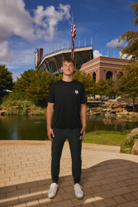 A man stands outdoors in front of a stadium and pond, wearing a black short-sleeve t-shirt with a small white logo on the left chest, black sweatpants, and white sneakers.