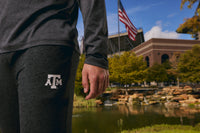 A close-up of a person wearing dark gray heather athletic pants with a white Texas A&M logo, standing outdoors near a pond and stadium in a real environment.