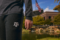 A close-up of a person wearing dark gray heather athletic pants with a white Texas A&M logo, standing outdoors near a pond and stadium in a real environment.