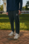 A person standing outdoors on a brick path in front of a grassy lawn and a classical building, wearing dark charcoal jogger sweatpants with a small white embroidered logo on the thigh, a navy long-sleeve top, and chunky off-white sneakers.