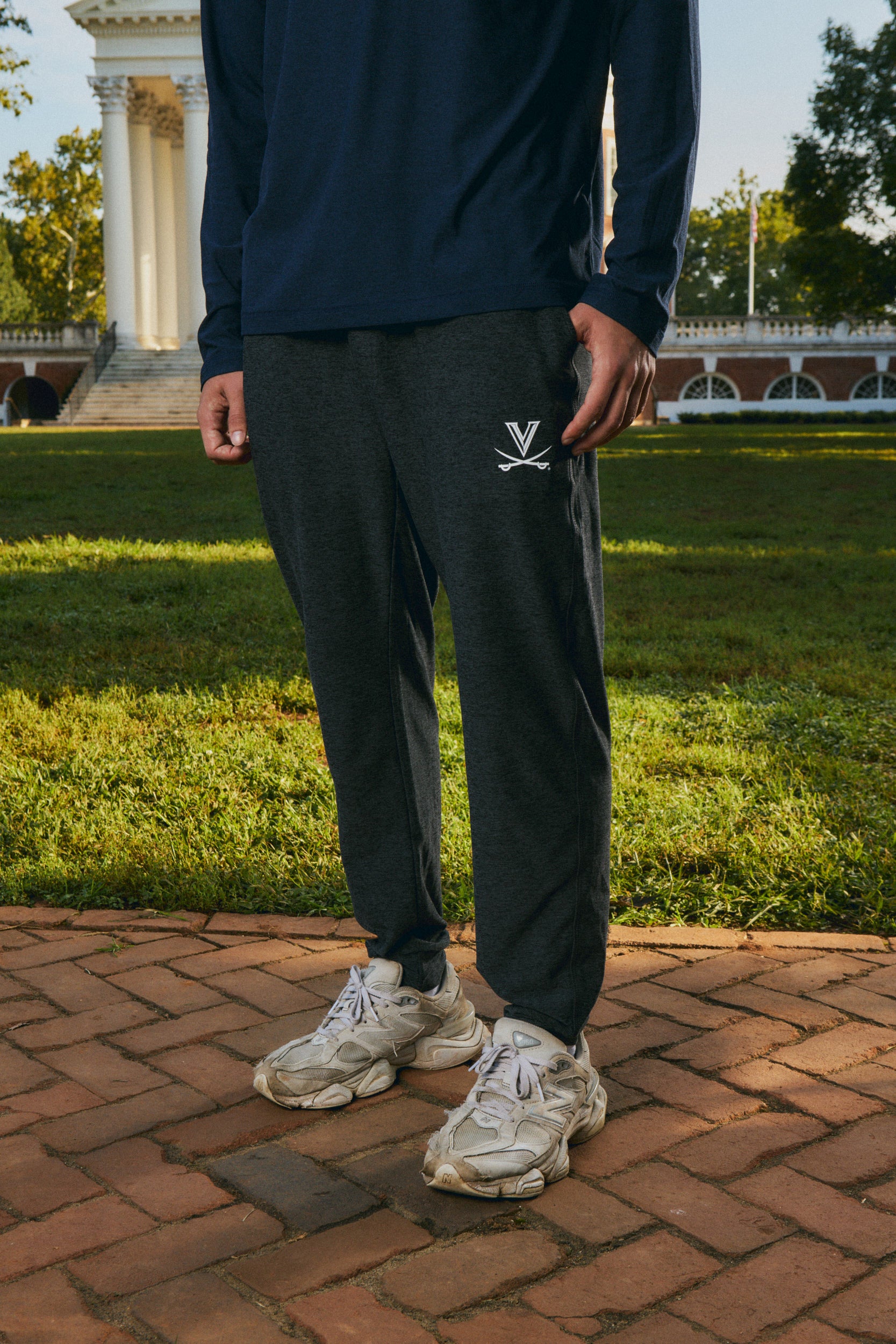 A person standing outdoors on a brick path in front of a grassy lawn and a classical building, wearing dark charcoal jogger sweatpants with a small white embroidered logo on the thigh, a navy long-sleeve top, and chunky off-white sneakers.
