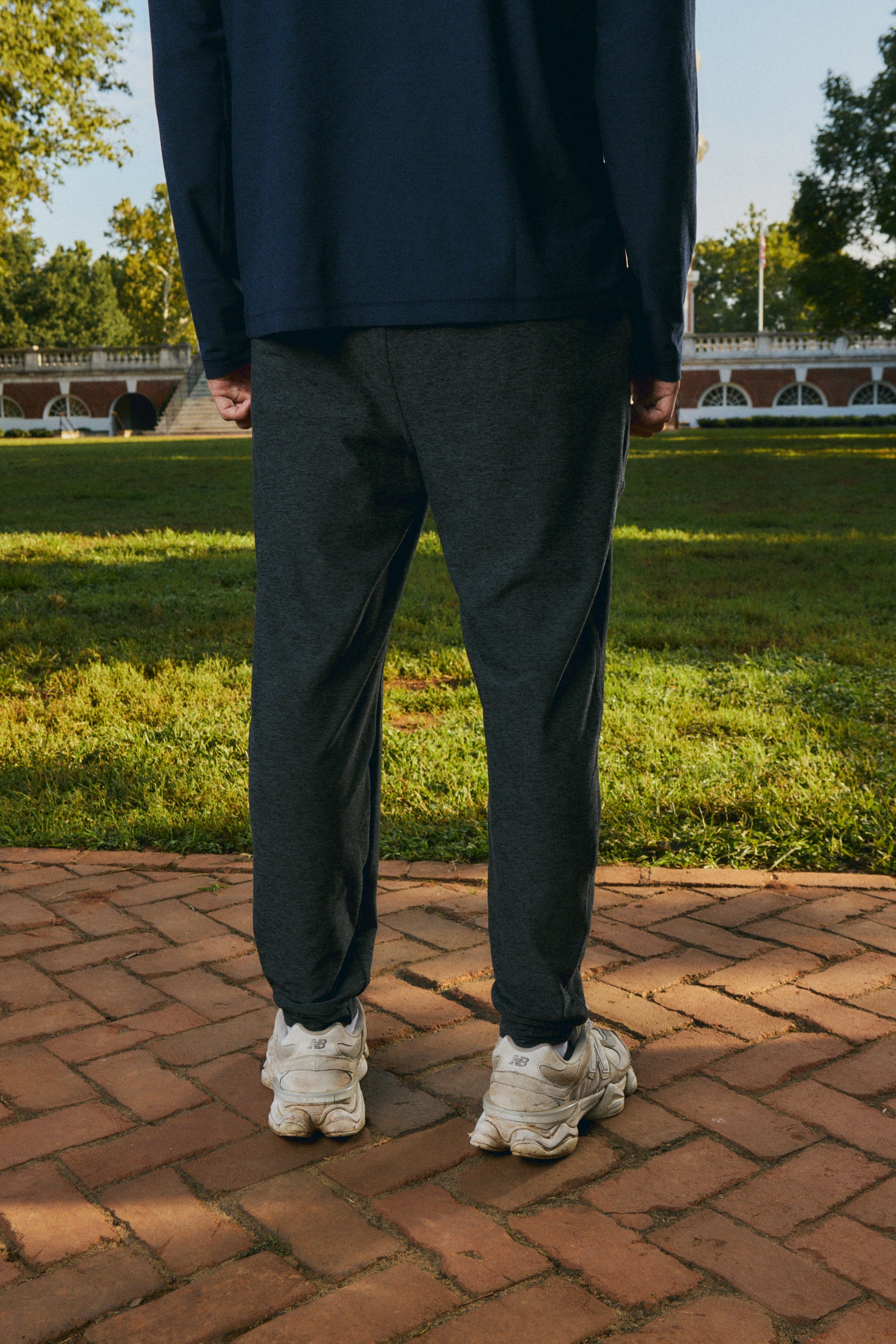 A person standing outdoors on a brick path in a park, wearing dark charcoal gray jogger pants, a navy long-sleeve top, and white sneakers.